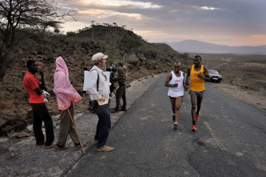 Eine asphaltierte Straße in Kenia führt direkt zum Betrachte und etwas bergan. Henry Wanyoike läuft diese in Tichtung Betrachter, links steht das Kamerateam. Schöne Landschaft leicht diesiges Abendlicht.