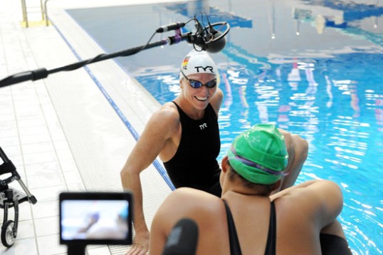 Vorne eine Schwimmerin, von der nur der Rücken zu sehen ist. Bildmittig Kirsten Brun in Schwimmzeug lachend, zuhörend. eim Mikrofon und ein Monitor ragten ins Bild.