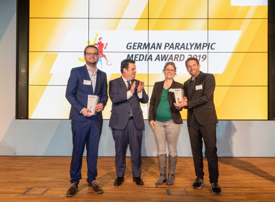 Das Foto zeigt die drei Preisträger mit Bundesminister Hubertus Heil vor einer Video-Wand. Der Minister applaudiert, die Preisträgerinnen halten den Preis in ihren Händen.