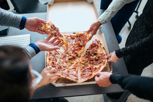 Ein Foto auf dem eine Pizza zu sehen ist. Mehrere Hände greifen nach der Pizza und nehmen sich ein Stück