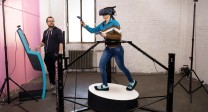 Foto: Versuchsaufbau: Frau mit VR-Brille und Handsensoren steht auf Aktionsplateau (3D-Laufband). Ein Mann steht beobachtend daneben.Bild: IFA
