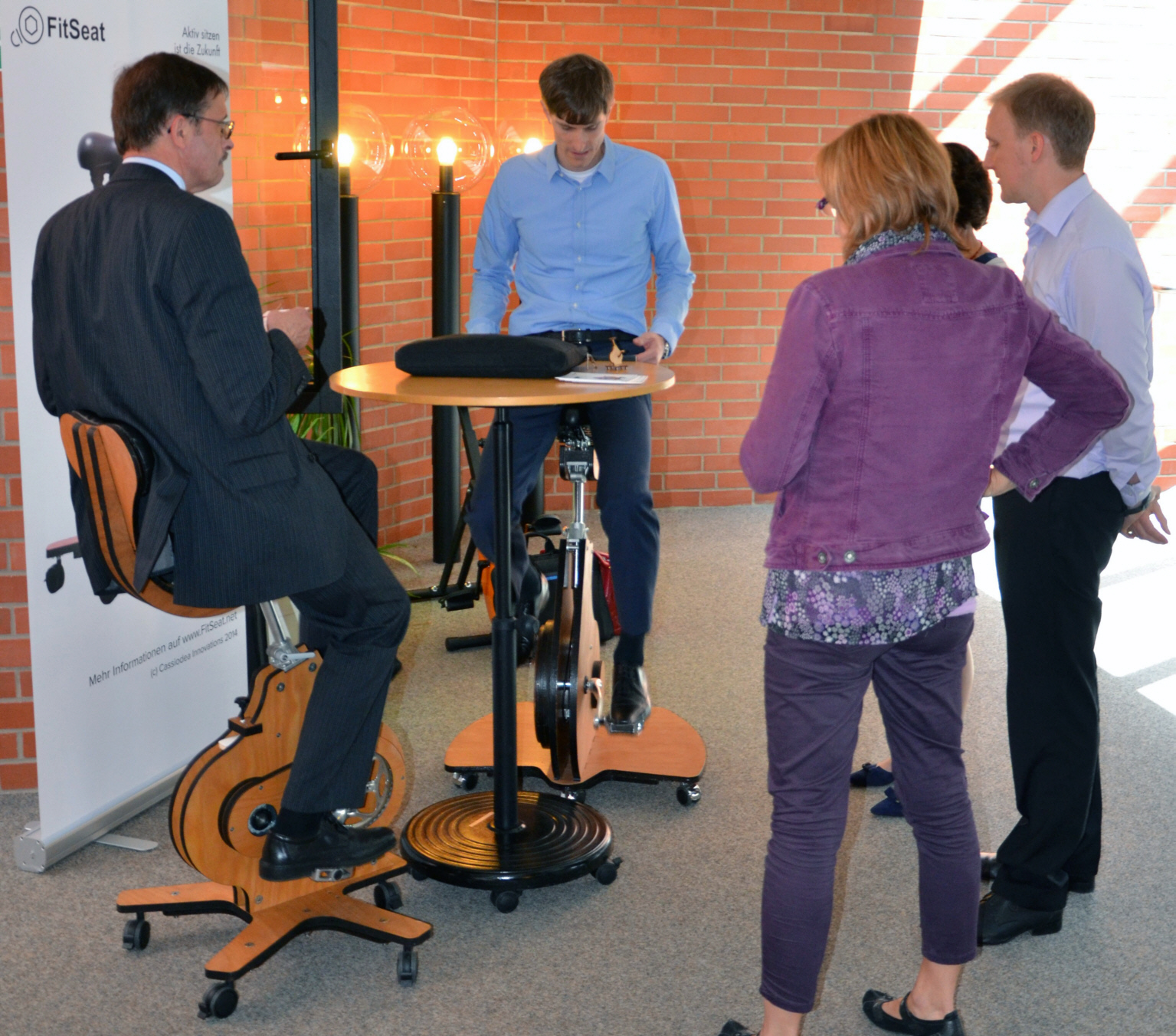 Two persons sitting at a dynamic workplace, three persons watching them