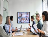 Personen sitzen in einem Büro und schauen auf einen Bildschirm mit einer Videokonferenz.