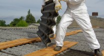 Zwei Personen in Schutzkleidung tragen eine Asbestplatte auf einem Dach.