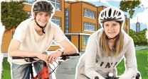 Zwei Jugendliche stehen mit ihren Fahrrädern vor einem modernen Schulgebäude. Beide tragen Fahrradhelme und lächeln in die Kamera.